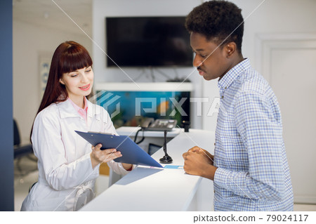 Young female nurse with clipboard showing application form to young male black patient at hospital Young female nurse with clipboard showing application form to young male black patient at hospital 79024117