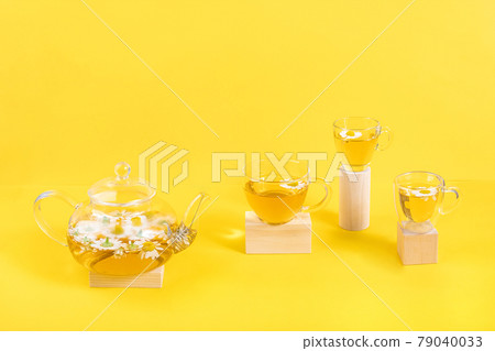 Three cups of camomile tea, transparent teapot on wood geometric shape, yellow background. Creative concept Natural Chamomile Tea. Top view Flat lay 79040033