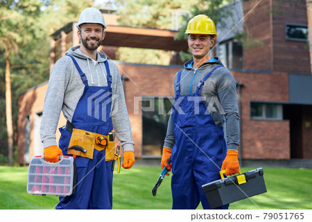 Rely on us. Two positive handsome young male engineers in hard hats smiling at camera, posing outdoors with toolbox and suitcase with tools while working on cottage construction site Rely on us. Two positive handsome young male engineers in hard hats smiling at camera, posing outdoors with toolbox and suitcase with tools while working on cottage construction site 79051765