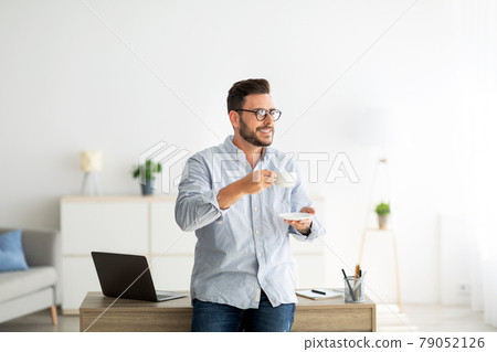 Young freelancer guy with cup of coffee standing at desk at home interior, leaning at table and looking away, copy space Young freelancer guy with cup of coffee standing at desk at home interior, leaning at table and looking away, copy space 79052126