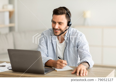 New knowledge online at home. Young man in headphones making notes and looking at laptop computer in living room New knowledge online at home. Young man in headphones making notes and looking at laptop computer in living room 79052127