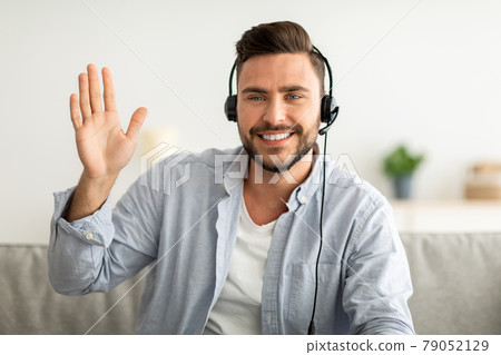 Online teacher's portrait. Happy handsome man wearing headset waving hand to webcam, gesturing hello Online teacher's portrait. Happy handsome man wearing headset waving hand to webcam, gesturing hello 79052129