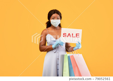 Black lady in mask and gloves showing SALE sign, holding shopper bags, buying goods on discount during coronavirus 79052561