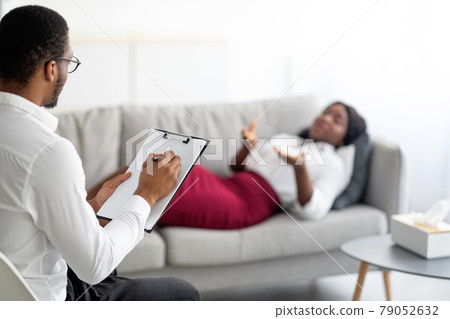 Young black woman on consultation with psychologist, lying on sofa at office, explaining about her problem to doctor 79052632