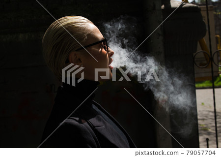 A beautiful girl smokes a vape and lets out smoke A beautiful girl smokes a vape and lets out smoke 79057704
