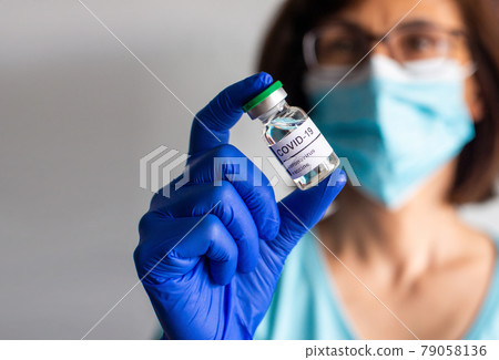 Middle age female nurse in safety mask and gloves holds bottle with liquid COVID vaccine for injections 79058136