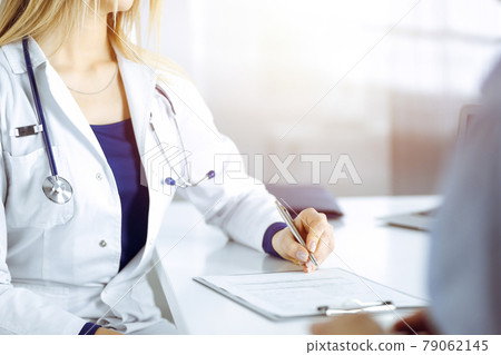 Unknown woman- doctor is listening to her patient, while sitting together at the desk in the sunny cabinet in a clinic. Female physician with a stethoscope is writing at clipboard, close up. Perfect Unknown woman- doctor is listening to her patient, while sitting together at the desk in the sunny cabinet in a clinic. Female physician with a stethoscope is writing at clipboard, close up. Perfect 79062145