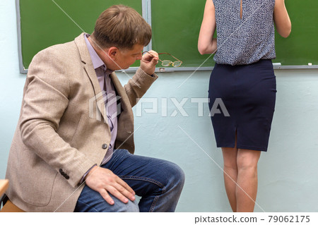 A male teacher watches a young attractive student at the blackboard through glasses 79062175