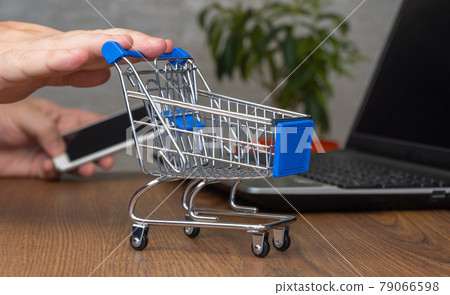 online shopping, concept of buying goods at home. miniature shopping cart. small supermarket trolley. man orders a product on a laptop and smartphone 79066598