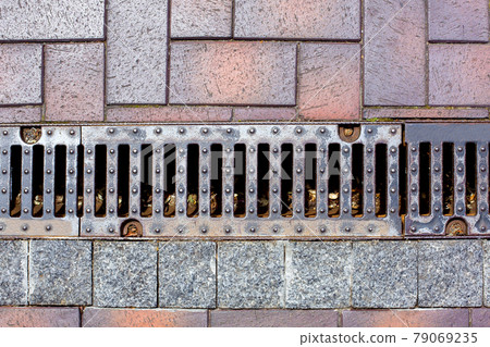 an iron drainage grate of a rain channel on a pedestrian sidewalk made of stone tiles urban landscaping infrastructure of parks. 79069235