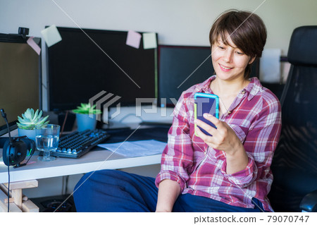 Smiling woman looking at the phone screen while sitting on her home office or office working place. Take a break. Reading the good news. Remote work, social networks, blogging. Telecommuting concept. Smiling woman looking at the phone screen while sitting on her home office or office working place. Take a break. Reading the good news. Remote work, social networks, blogging. Telecommuting concept. 79070747