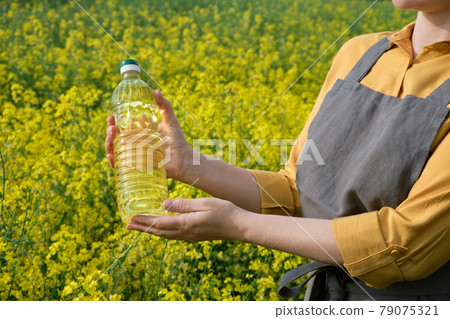 A woman stands in a rape field and holds a bottle of rapeseed oil in his hands. Concept of canola oil production. A woman stands in a rape field and holds a bottle of rapeseed oil in his hands. Concept of canola oil production. 79075321