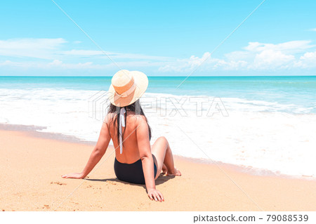 Back view of asian traveler woman in swimsuit and straw hat to sitting in the spray of the sea to looking view 79088539