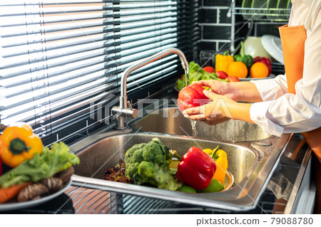 Asian housewife wearing an apron and standing to washing bell pepper and vegetables with water in sink Asian housewife wearing an apron and standing to washing bell pepper and vegetables with water in sink 79088780