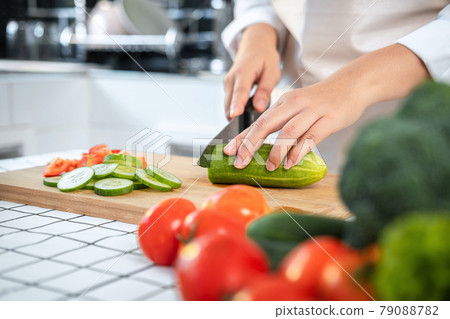 Close up of asian housewife wearing apron and using knife to slice cucumber and tomato on chopping board Close up of asian housewife wearing apron and using knife to slice cucumber and tomato on chopping board 79088782