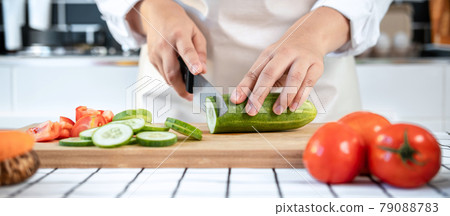 Close up of asian housewife wearing apron and using knife to slice cucumber and tomato on chopping board Close up of asian housewife wearing apron and using knife to slice cucumber and tomato on chopping board 79088783