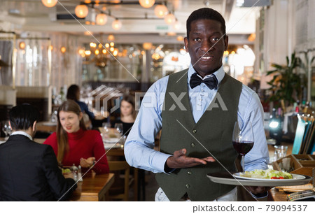 Man waiter is holding tray with order in hall of restaurant. Man waiter is holding tray with order in hall of restaurant. 79094537