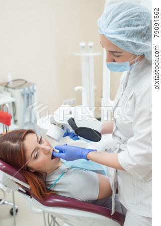 Attractive smiling woman doctor in a white uniform with tools in hand. Young beautiful female dentist takes jaw x-ray of female patient. Attractive woman patient at dentist taking x-ray exam. 79094862
