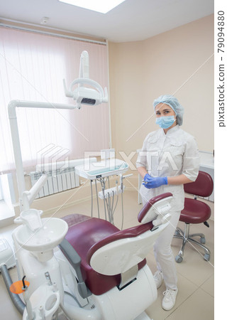 Girl dentist, in white clothes, mask and cap stands next to a modern dental chair in a large, simple, medical office Girl dentist, in white clothes, mask and cap stands next to a modern dental chair in a large, simple, medical office 79094880