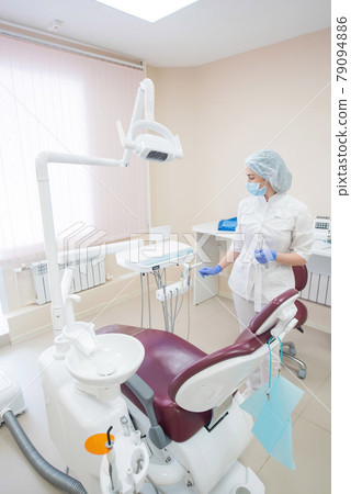 Girl dentist, in white clothes, mask and cap stands next to a modern dental chair in a large, simple, medical office Girl dentist, in white clothes, mask and cap stands next to a modern dental chair in a large, simple, medical office 79094886