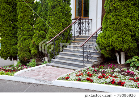 a granite staircase with a wrought iron railings, entrance steps, exit to the backyard with a flowerbed with flowers and evergreen thuja trees, nobody. 79096926