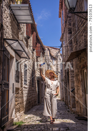 Beautiful blonde young female traveler wearing straw sun hat, taking selfie while sightseeing and enjoying summer vacation in an old traditional costal town at Adriatic cost, Croatia. 79103613