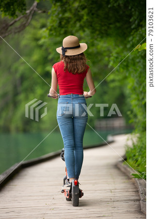 Rear view of casual female tourist riding urban electric scooter in beautiful natural enviorment by lake Bled, Slovenia. Rear view of casual female tourist riding urban electric scooter in beautiful natural enviorment by lake Bled, Slovenia. 79103621