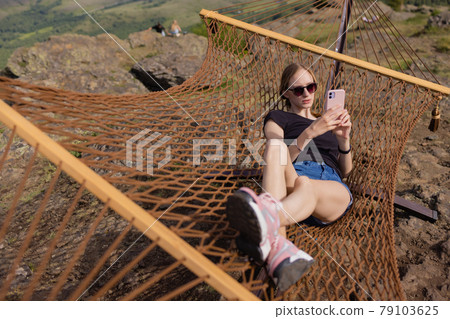 Young adult happy short haired shaved bald attractive woman chilling enjoy relaxing sitting in fabric chair hammock at yard in garen near house. Peaceful and idyllic outdoor rest lifestyle. 79103625