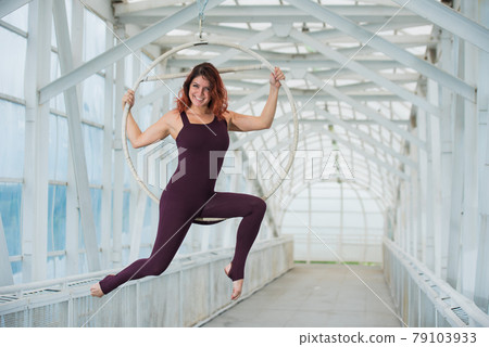 The red-haired aerial gymnast in a sports combo performs at the air hoop in a pedestrian crossing. 79103933