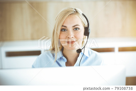 Blonde business woman sitting and communicated by headset in call center in sunny office. Concept of telesales business 79108738