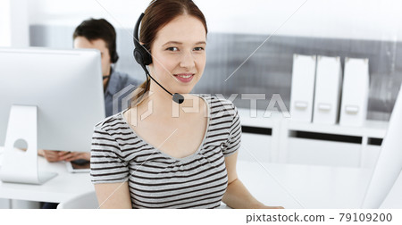 Casual dressed young woman using headset and computer while talking with customers online. Group of operators at work. Call center, business concept Casual dressed young woman using headset and computer while talking with customers online. Group of operators at work. Call center, business concept 79109200
