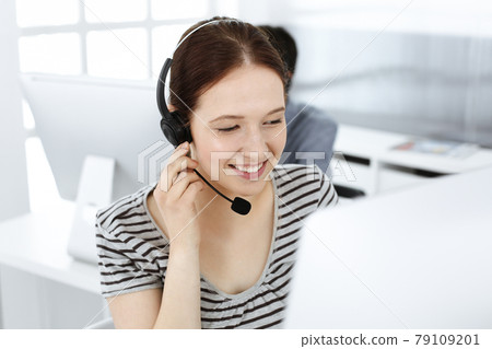 Casual dressed young woman using headset and computer while talking with customers online. Group of operators at work. Call center, business concept Casual dressed young woman using headset and computer while talking with customers online. Group of operators at work. Call center, business concept 79109201