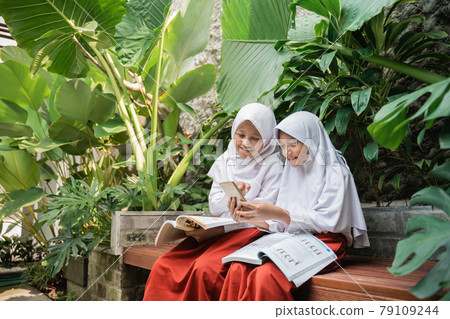 two children wearing veil in school uniforms using a cellphone and a book while studying together two children wearing veil in school uniforms using a cellphone and a book while studying together 79109244
