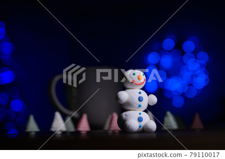 Close-up of a sweet snowman from a pastry mastic on a background of marshmallow trees and a white mug with cocoa. Candies in the dark against the background of a Christmas tree in the lights. Close-up of a sweet snowman from a pastry mastic on a background of marshmallow trees and a white mug with cocoa. Candies in the dark against the background of a Christmas tree in the lights. 79110017