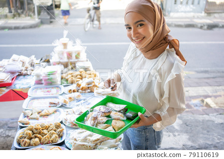 A beautiful girl wearing a headscarf uses tongs and carrying a plastic tray choosing snacks A beautiful girl wearing a headscarf uses tongs and carrying a plastic tray choosing snacks 79110619