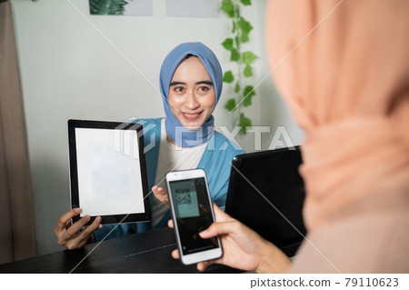 A hijab business woman holds a tablet and shows the screen to customers who use cellphones while paying non-cash by scanning barcodes A hijab business woman holds a tablet and shows the screen to customers who use cellphones while paying non-cash by scanning barcodes 79110623