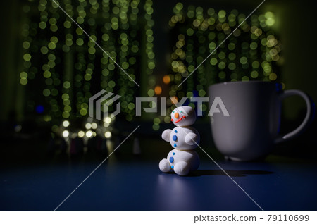 Cute snowman and little Christmas trees made of marshmallows on the background of Christmas lights. White mug of hot chocolate and sweets. 79110699