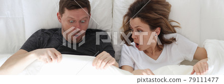 Surprised man and woman lying in bed and looking under covers top view Surprised man and woman lying in bed and looking under covers top view 79111477