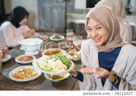 A veiled woman smiles at the camera with hand gestures serving while holding a plate of vegetables 79113827