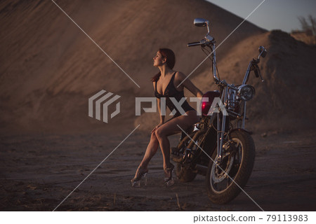 Red-haired woman in sexy lingerie in high heels sits on a motorcycle. Attractive red-haired girl sits on a motorcycle at sunset 79113983