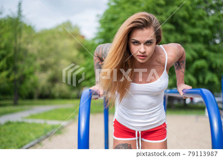 Athletic woman doing triceps exercises on the uneven bars on the playground on a hot summer day. Female bodybuilder in shorts and a short top trains outdoors during quarantine. Embossed Muscular body. Athletic woman doing triceps exercises on the uneven bars on the playground on a hot summer day. Female bodybuilder in shorts and a short top trains outdoors during quarantine. Embossed Muscular body. 79113987