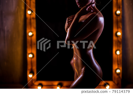 A young, naked, Caucasian girl with a very slender figure poses against a large mirror with lights in the loft style. The skin is oiled and shiny. Sexy tan. A young, naked, Caucasian girl with a very slender figure poses against a large mirror with lights in the loft style. The skin is oiled and shiny. Sexy tan. 79114379