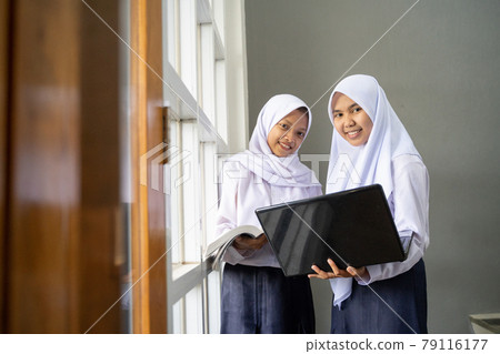 Two Asian teenagers wearing headscarves in school uniforms carry a laptop with copyspace 79116177