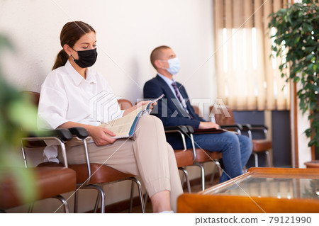 Concentrated girl in a protective mask, sitting in the lobby looks at an information magazine 79121990