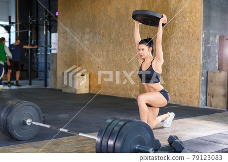 Woman during training in the gym. She lunges and holds the weight above her head. 79123083