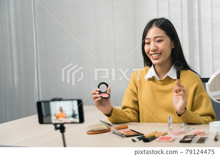 Young asian woman vlogging about cosmetics skin care items products on table with her video camera and demonstrates product use and reviews for her online blog channel. Young asian woman vlogging about cosmetics skin care items products on table with her video camera and demonstrates product use and reviews for her online blog channel. 79124475