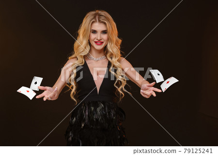 Blonde girl in black dress and necklace. She is smiling, holding or throwing something, posing on brown background. Template or mockup. Close up Blonde girl in black dress and necklace. She is smiling, holding or throwing something, posing on brown background. Template or mockup. Close up 79125241