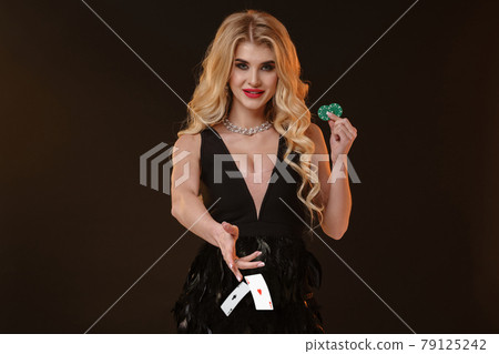 Blonde model in black dress and necklace. Smiling, showing two green chips, throwing something, posing on brown background. Poker, casino. Close-up Blonde model in black dress and necklace. Smiling, showing two green chips, throwing something, posing on brown background. Poker, casino. Close-up 79125242