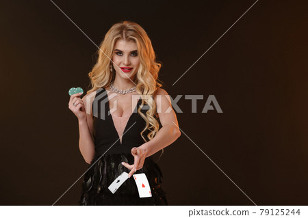 Blonde girl in black dress and necklace. Smiling, showing two green chips, throwing something, posing on brown background. Poker, casino. Close-up Blonde girl in black dress and necklace. Smiling, showing two green chips, throwing something, posing on brown background. Poker, casino. Close-up 79125244