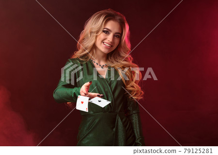Blonde girl in green stylish dress and jewelry. She is smiling and reaching her hand to you, posing against colorful smoky background. Close-up. 79125281